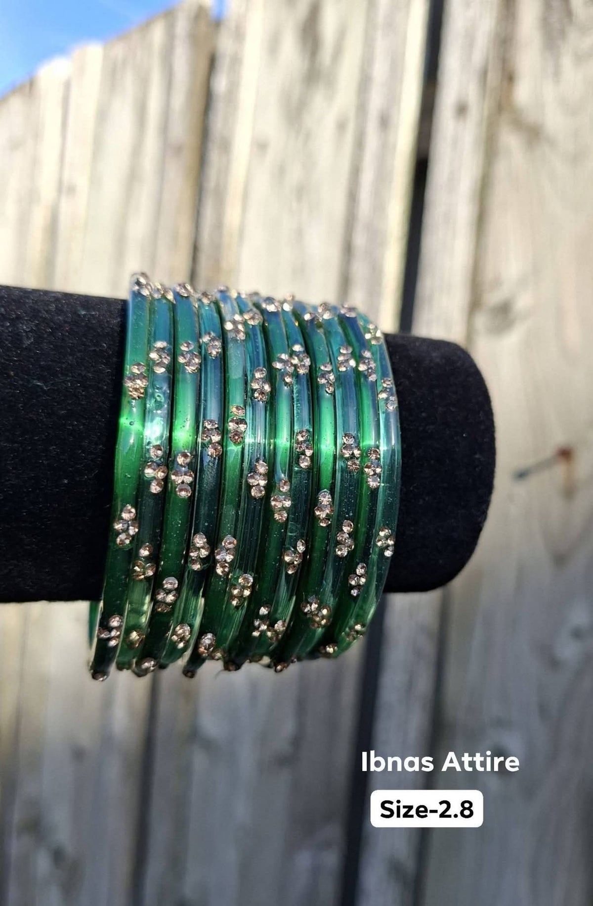 Set of 12 translucent emerald green glass bangles studded with golden rhinestone dots displayed on a black velvet display stand outdoor against a wooden fence