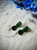 Silver-tone drop earrings featuring a rectangular emerald green stone linked to a teardrop-shaped green stone, laid flat on a white fabric surface with blue floral background elements.