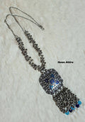 Long oxidized silver chain necklace with clustered bead links, a square blue enamel pendant, and ghungroo tassel drops with blue glass beads displayed on a white faux fur surface.  
