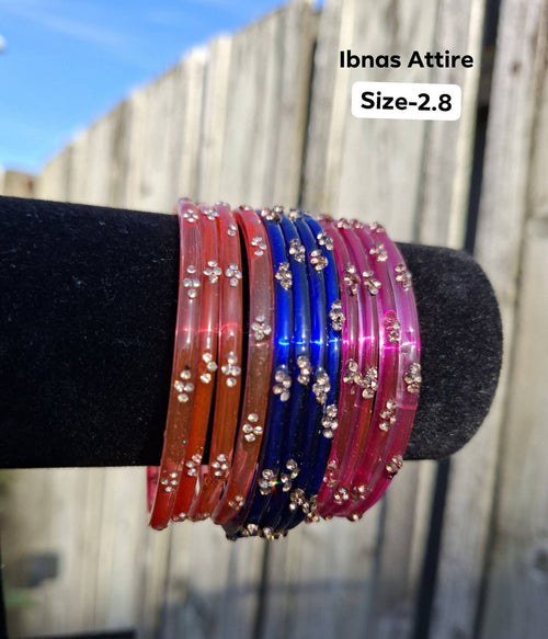 Stack of translucent glass bangles in coral orange, royal blue, and magenta pink with gold sequin accents displayed on a black velvet bar outdoors against a wooden fence.